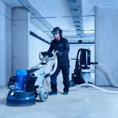 Man i skyddsutrustning slipar betonggolv med en maskin. En dammsugare är ansluten för att samla upp damm.