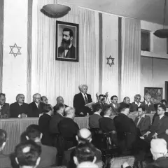 Hombres en traje asisten a un evento histórico, la creacion del estado de Israel, con la bandera israelí y un retrato de Theodor Herzl.