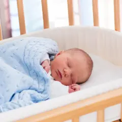 Śpiące niemowlę w łóżeczku, otulone błękitnym kocykiem.