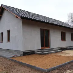 Nowy dom w trakcie budowy, z szarym tynkiem i brązowymi oknami. Wniosek o warunki zabudowy złożony, teren wokół domu przygotowany do dalszych prac.