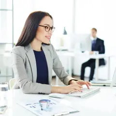 Kobieta w okularach pracuje przy komputerze, analizując raporty. W tle inni pracownicy, co sugeruje podwyższone koszty uzyskania przychodu w firmie.