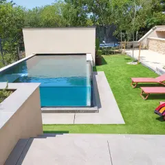Un magnifique bassin vitré aux eaux bleues, entouré d'une pelouse synthétique et de chaises longues roses.