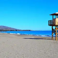 Torre de vigilancia en la vera playa, con el mar azul y montañas al fondo.