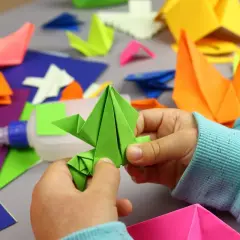 Dziecko tworzy proste origami z kartki a4. Wokół kolorowe figurki i klej.