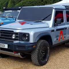 Srebrny INEOS Grenadier z czerwonym logo "A" na drzwiach, gotowy na przygodę. Obok niebieski SUV.