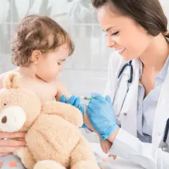 Lekarka podaje dziecku szczepionkę MMR. Maluch trzyma misia, a pielęgniarka przytrzymuje jego ramię.