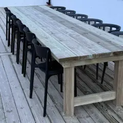 Ett stadigt, hemmabyggt utebord av trä med plats för många gäster. Svarta stolar står uppradade runt det robusta bordet på altanen.