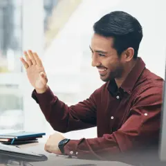 Man waves hello during an online emotional intelligence coaching session, smiling warmly at his laptop.