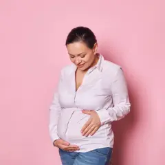 Uśmiechnięta kobieta w ciąży po 40. delikatnie dotyka brzucha na różowym tle.