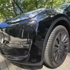 Błyszczący, czarny samochód elektryczny zaparkowany na chodniku z kostki brukowej, w tle drzewa i budynek.