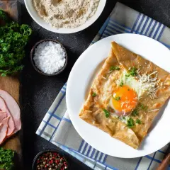 Francuska crepe z jajkiem sadzonym, serem i natką pietruszki na talerzu, obok szynka i składniki.