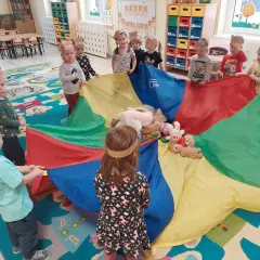 Dzieci w opaskach z uszami misia bawią się kolorowym spadochronem z pluszakami.