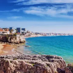 Felsige Küste mit Sandstrand und weißen Häusern, Blick auf das türkisfarbene Meer und die Skyline von Lissabon. Ein perfekter lissabon strand.
