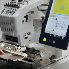Une machine à broder professionnelle, type impression DTG, crée un logo sur un tissu. L'écran tactile affiche les détails du projet.