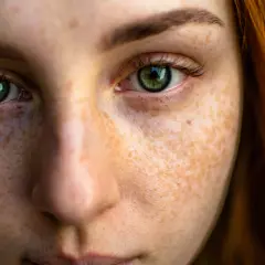 Close-up texture photography of a young woman's face, highlighting her green eyes and the intricate pattern of her freckles.