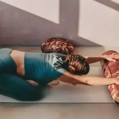 Kobieta w pozycji balasana na macie do jogi, z rękami wyciągniętymi do przodu i podpartymi wałkiem.