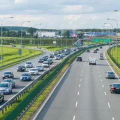 Autostrada z ruchem samochodowym w słoneczny dzień, z zielonymi poboczami i drzewami w tle.