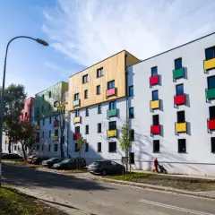 Nowoczesne mieszkanie TBS z kolorowymi balkonami i drewnianymi elementami fasady, zaparkowane samochody i osoba pchająca wózek.
