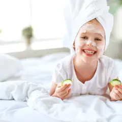 Uśmiechnięta dziewczynka z ręcznikiem na głowie i białą glinką na twarzy cieszy się domowym spa, trzymając w dłoniach plasterki ogórka.