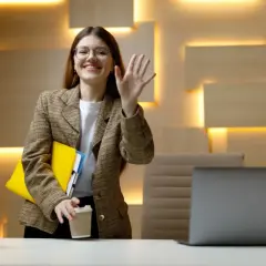 Uśmiechnięta kobieta w okularach, z teczkami i kawą, macha ręką. To jej dzień próbny w pracy bez umowy, pełen nadziei.