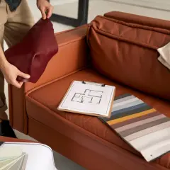 A person is choosing couch materials, holding a burgundy swatch next to a brown leather couch. Fabric samples and a floor plan are on the seat.
