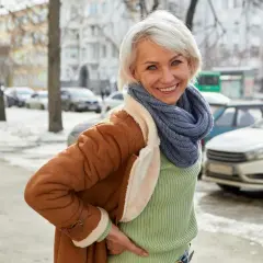 Uśmiechnięta kobieta w brązowym kożuchu i niebieskim szaliku stoi na tle zimowego miasta.