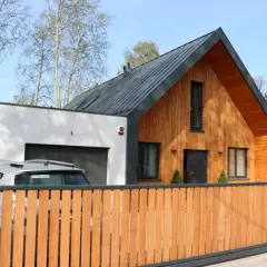 Nowoczesny dom z murowaną częścią i drewnianą dobudówką. Obok garaż, przed którym stoi samochód.