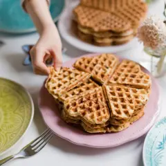 Ein Kind greift nach einem herzförmigen Waffel ohne Ei. Mehrere Stapel Waffeln liegen auf rosa und weißen Tellern, umgeben von bunten Tellern und Blumen.