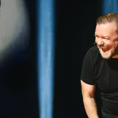 Ricky Gervais w swoim stand upie śmieje się do rozpuku, w czarnym t-shircie, na tle niebieskich świateł.