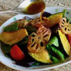 A spoon pours a golden dressing over a salad of roasted pumpkin, lotus root, tomatoes, and greens. The dressing, with a hint of wasabi, adds a zesty kick.