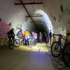 Grupa rowerzystów eksploruje mroczną, graffiti-pokrytą podziemną trasę rowerową, oświetloną latarkami.