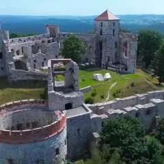 Ruiny zamku Tenczyn na wzgórzu, otoczone zielenią. Widać wieże, mury obronne i dziedziniec z namiotami.