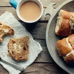 Na drewnianym stole leżą świeże, pachnące hot cross buns, obok kubek kawy i kromka posmarowana masłem.