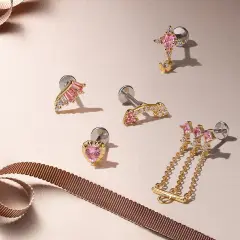 Złote kolczyki do ucha, w tym subtelny tragus piercing w kształcie serca i skrzydła, ozdobione różowymi kamieniami i cyrkoniami.