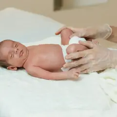 Niemowlę z lekką plagiocefalią leży na białym ręczniku, a ręce w rękawiczkach delikatnie dotykają jego nóżek.