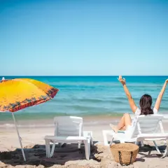 Kobieta cieszy się urlopem wypoczynkowym na plaży, z rękami w górze. Obok leżaki i parasol.