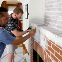Two men are learning how to whitewash brick on a fireplace, applying white paint with brushes for a rustic look.