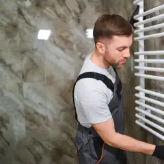 Un plombier en salopette ajuste la vanne d'un sèche serviette blanc, prêt pour son installation.