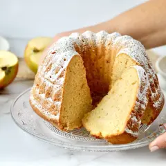 Ein saftiger Kuchen mit Apfelmus, bestäubt mit Puderzucker. Daneben liegen Äpfel und ein Glas Apfelmus.