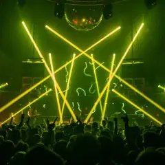 Folla in delirio in un techno club, illuminato da fasci di luce gialla e una sfera stroboscopica.