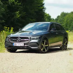 Szary Mercedes-Benz Klasy C All-Terrain na szutrowej drodze, otoczony zielenią lasu i polnymi kwiatami.
