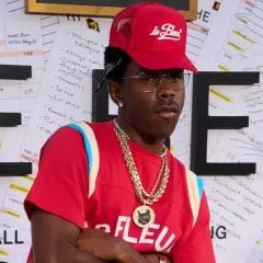Tyler, The Creator w czerwonej czapce i naszyjnikach, na tle liter "HE" i notatek.