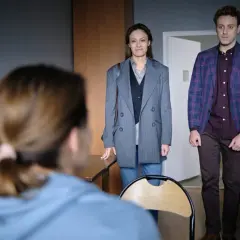 Dwoje ludzi stoi w pokoju, patrząc na trzecią osobę zza kadru.