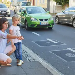 Mutter erklärt ihrer Tochter, dass sie auf dem Gehweg nicht in falsche Richtung fahren darf. Ein grünes Auto fährt auf der Straße.