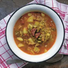 Gęsta zupa z kapusty włoskiej z ziemniakami i kiełbasą, podana w białej misce na kratkowanej serwetce.