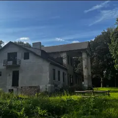 Zniszczony dworek z kolumnami i balkonem, otoczony zielenią.