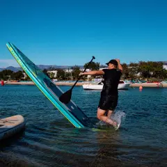 Mężczyzna w wodzie próbuje utrzymać równowagę na desce podczas pływania na supie. W tle widać plażę i inne deski.