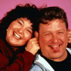 Roseanne und Dan, die Hauptfiguren der besetzung von Roseanne, lachen und Grimassen vor einem rosa Hintergrund.