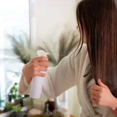 Kobieta w szlafroku spryskuje włosy odżywką w sprayu.