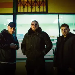 Trzech mężczyzn w ciemnych kurtkach, tworzących zgraną hałastrę, pozuje przed oświetloną witryną sklepową.